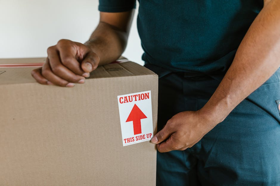 Close-up view of a person wearing a dark shirt and blue work trousers, holding a large cardboard moving box during a house relocation. The individual is applying a caution label with a red upward arrow and the instructions 'THIS SIDE UP' to the top side of the box, which is inside a well-lit room. The background shows a plain wall and a piece of furniture or shelving unit, indicating an indoor environment. The box is surrounded by other packing materials, and the person appears to be preparing it for safe transport. This scene illustrates the careful packing and handling procedures involved in home removals and furniture transport services provided by Man and a Van Finchley, aligning with their house removals guide for tight moves on Ballards Lane Finchley Central.