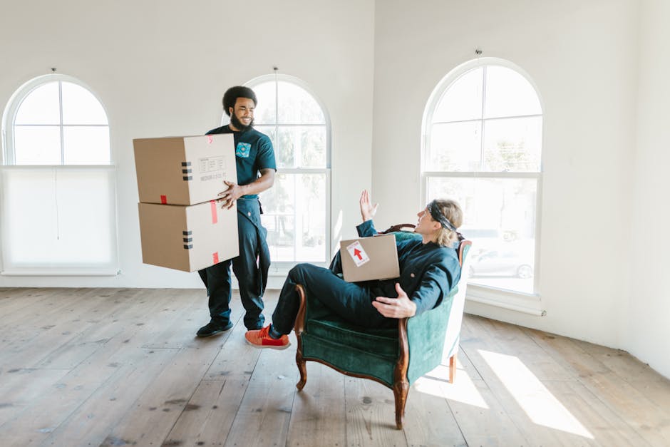 Close-up view of a person wearing a dark shirt and blue work trousers, holding a large cardboard moving box during a house relocation. The individual is applying a caution label with a red upward arrow and the instructions 'THIS SIDE UP' to the top side of the box, which is inside a well-lit room. The background shows a plain wall and a piece of furniture or shelving unit, indicating an indoor environment. The box is surrounded by other packing materials, and the person appears to be preparing it for safe transport. This scene illustrates the careful packing and handling procedures involved in home removals and furniture transport services provided by Man and a Van Finchley, aligning with their house removals guide for tight moves on Ballards Lane Finchley Central.