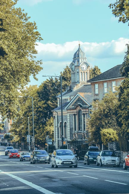 The image depicts a street scene in a residential area with several parked cars along the curb, including sedans and hatchbacks, in front of a historic church building with a prominent clock tower topped by a pointed spire. The church features arched windows, decorative brickwork, and a small entrance porch, surrounded by lush green trees that partially obscure some of the building's facade. The sky is bright with some scattered clouds, and sunlight casts shadows on the pavement and vehicles. In the background, there are additional residential buildings visible behind the trees. The scene suggests a peaceful neighborhood likely involved in home relocations or moving services, with possible activity related to loading or unloading furniture and boxes nearby, as indicated by the context of house removals. The image aligns with household transport and logistics services offered by Man and a Van Finchley, reflecting the process of moving or relocating within or around Finchley.