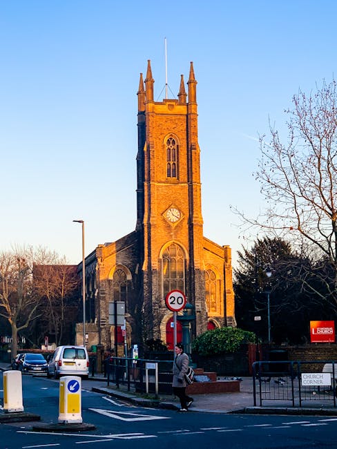 A tall stone church steeple with Gothic-style arched windows and decorative battlements rises above surrounding trees with green foliage in a clear sky. The church, set within a residential area, features a weathered stone exterior with narrow vertical windows on its tower. To the left, large leafy trees partially obscure the lower part of the structure, while on the right, bare branches are visible against the blue sky. The scene is illuminated by natural daylight, highlighting the texture of the stonework. This exterior view of the historic church contrasts with the nearby greenery, exemplifying a typical setting for house removals and furniture transport services provided by Man and a Van Finchley, supporting reliable home relocation logistics near Finchley Church End N12.