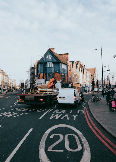 A white flatbed truck from Man and a Van Finchley is parked in the middle of a busy street in North Finchley, near the Tally Ho area, during daytime. The truck is loaded with stacks of cardboard boxes, wooden furniture, and packing materials, including plastic wrap and protective blankets, ready for home relocation or furniture transport. The truck is positioned across the lane, and part of the vehicle is aligned with the kerb, while the front faces an intersection with traffic lights. Behind the truck, there is a blue and brick residential building with multiple floors and windows, and further along the street, other buildings, street lamps, bicycles, and pedestrians are visible. The street features clear road markings, including a 20 mph speed limit circle, and the weather appears partly cloudy with natural daylight illuminating the scene, illustrating typical logistics involved in house removals or moving services in the area.