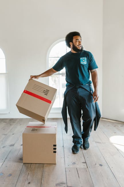 A man with a dark beard and curly black hair, dressed in a navy blue t-shirt with a light blue logo on the chest and dark trousers, is seen inside a bright, empty room with white walls and wooden flooring. He is holding a cardboard box sealed with red tape in his right hand while standing next to two other sealed cardboard boxes placed on the floor, with one slightly stacked on top of the other. The scene suggests he is engaged in packing or preparing items for a home relocation, fitting within a house removal or furniture transport process. Natural light streams through a large, arched window in the background, illuminating the space, and the room appears to be in a residential property. This image reflects the packing aspect of house removals and illustrates the careful handling of boxes typical of professional moving services such as those offered by Man and a Van Finchley.